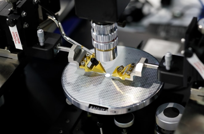 A semiconductor wafer under a light scope with two gold pinchers pointing at one chip on the wafer